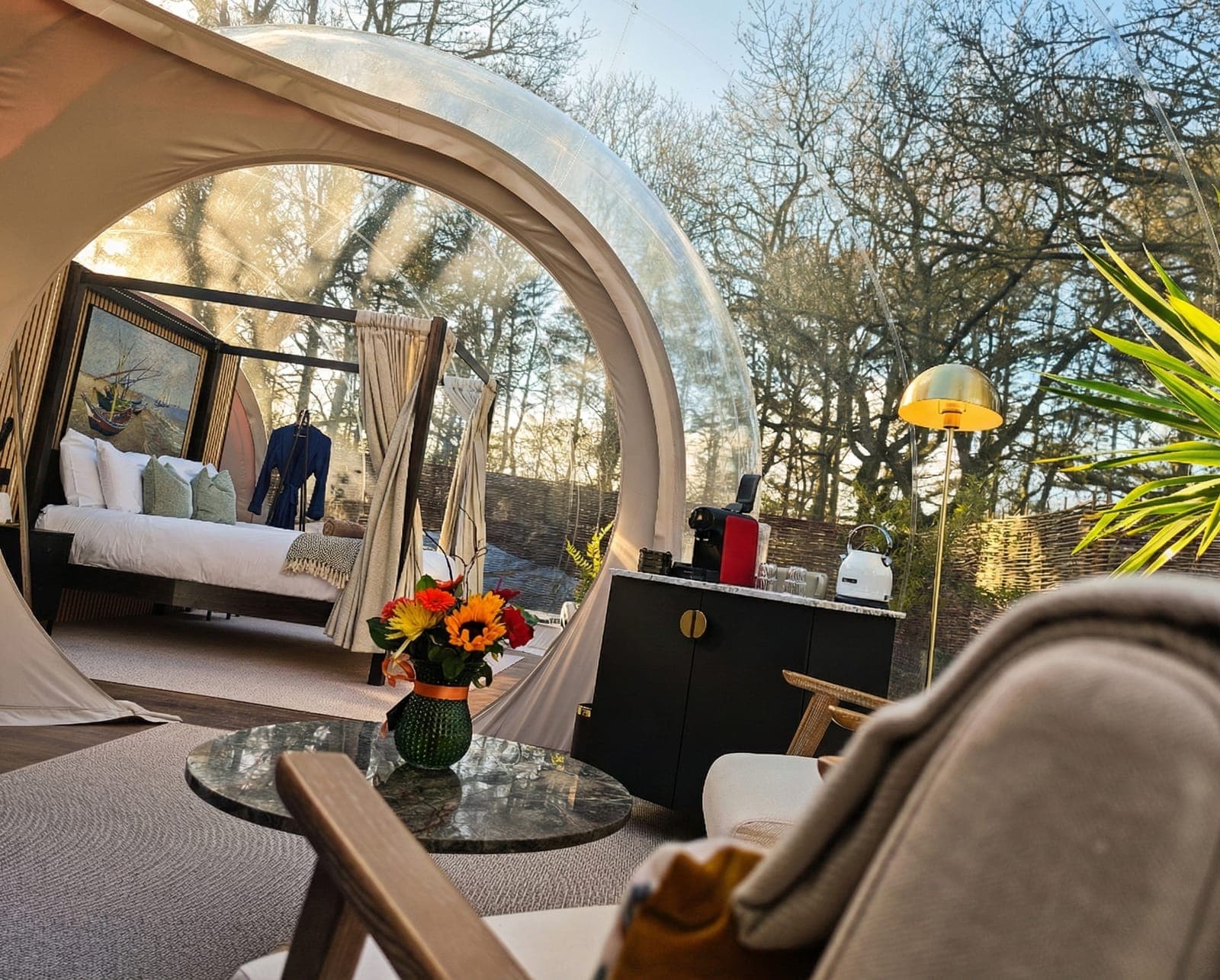 Inside a bubble dome at Cromore Retreat — four-poster bed, marble table with fresh flowers, and woodland light through the transparent walls