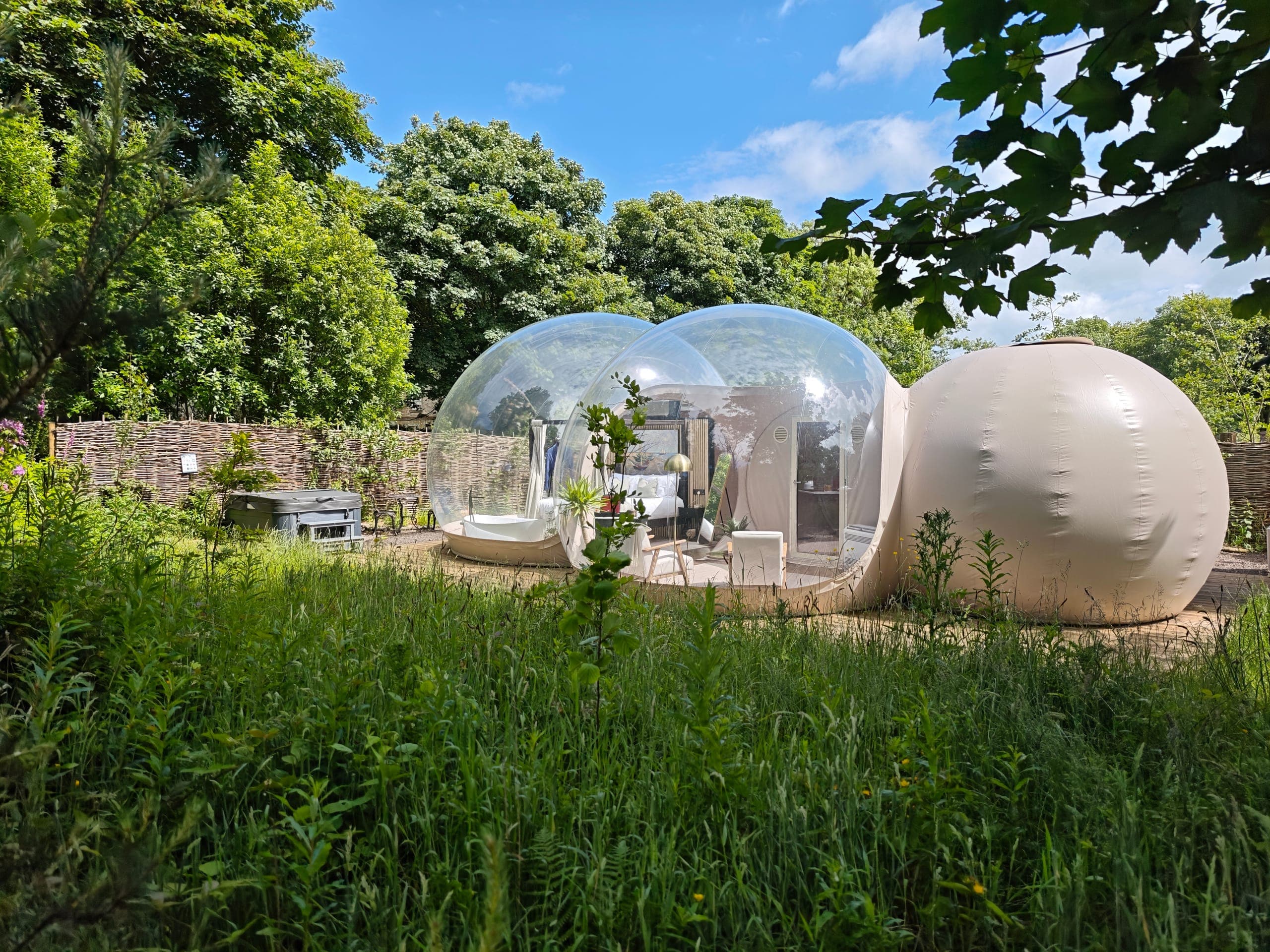 Bubble dome suite at Cromore Retreat, Portstewart — transparent domes with a four-poster bed and freestanding bath, set in wildflower meadow under a blue summer sky