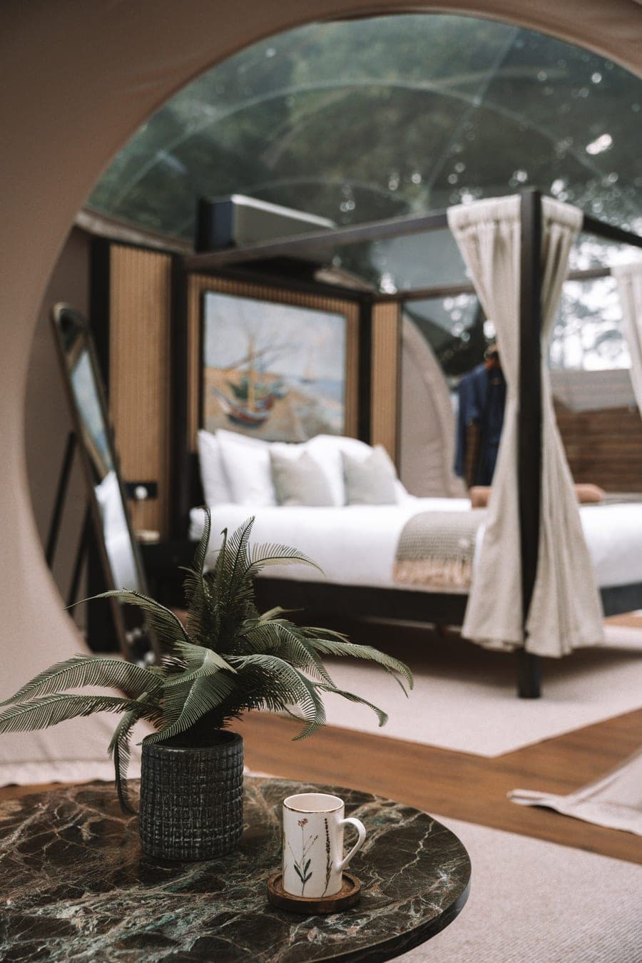 Inside the dome — four-poster bed beyond a marble table, framed by the transparent roof and the trees above