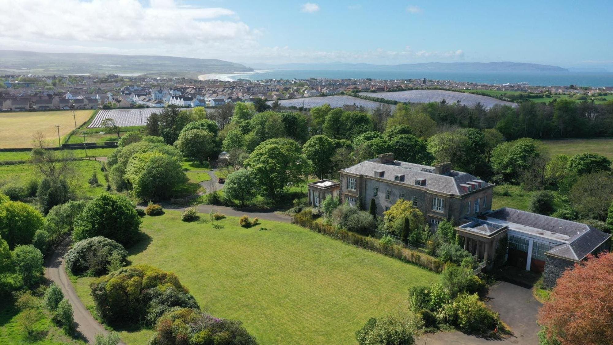 Aerial view of the Cromore Estate in Portstewart — the historic house and wooded grounds with the domes among the trees, and Portstewart Strand and the open Atlantic beyond